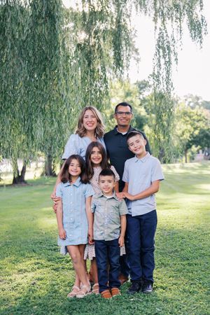 A family group poses together on a grassy hill under drooping willow trees in coordinated casual outfits.