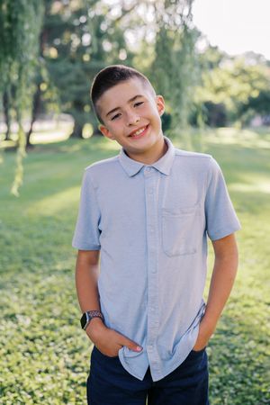 A young person in a light blue button-down shirt and dark pants poses playfully in a sunny outdoor setting.