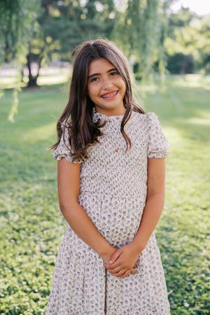 A person in a floral dress poses playfully in a sunny park setting with willow trees in the background.