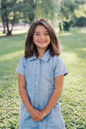 A child wearing a denim dress smiles while standing in a green outdoor setting.