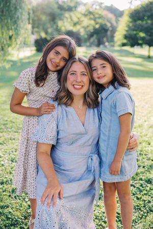 Three people in casual summer outfits pose together outdoors in a sunny field with greenery in the background.