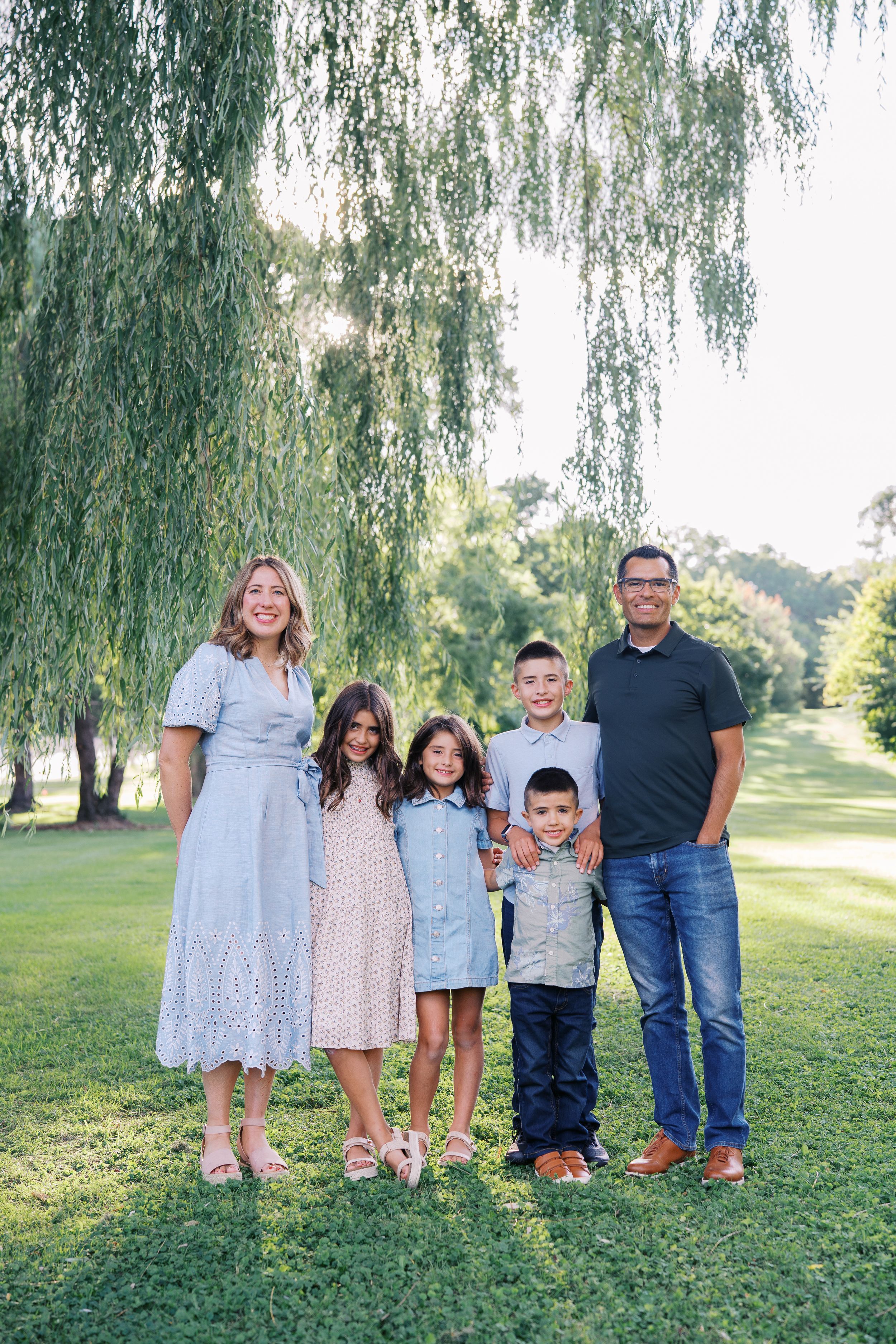 A family group poses together on grass under a weeping willow tree in coordinated blue and white outfits.