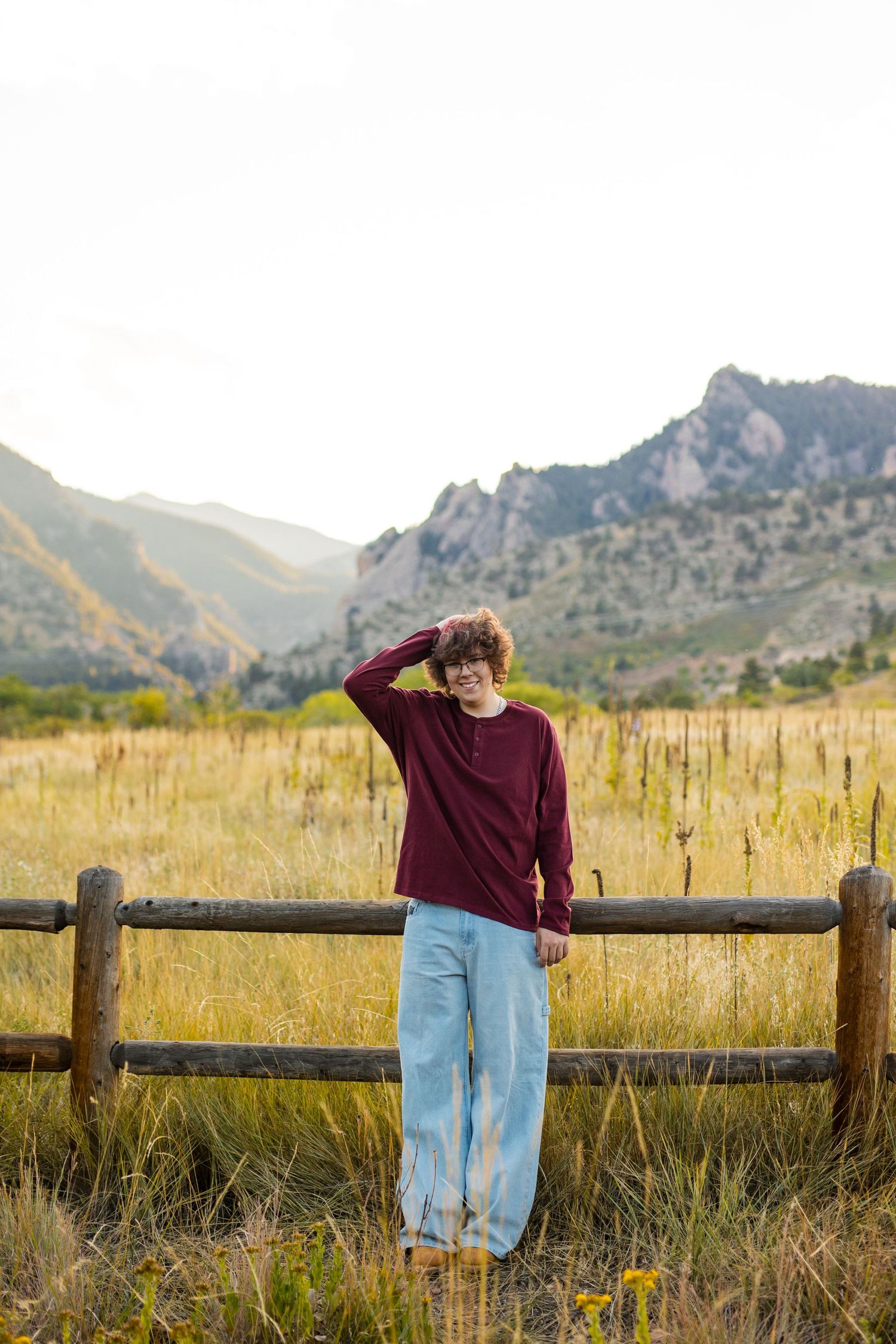 Person in burgundy sweater and light jeans stands by wooden fence with scenic mountain landscape in background at sunset.