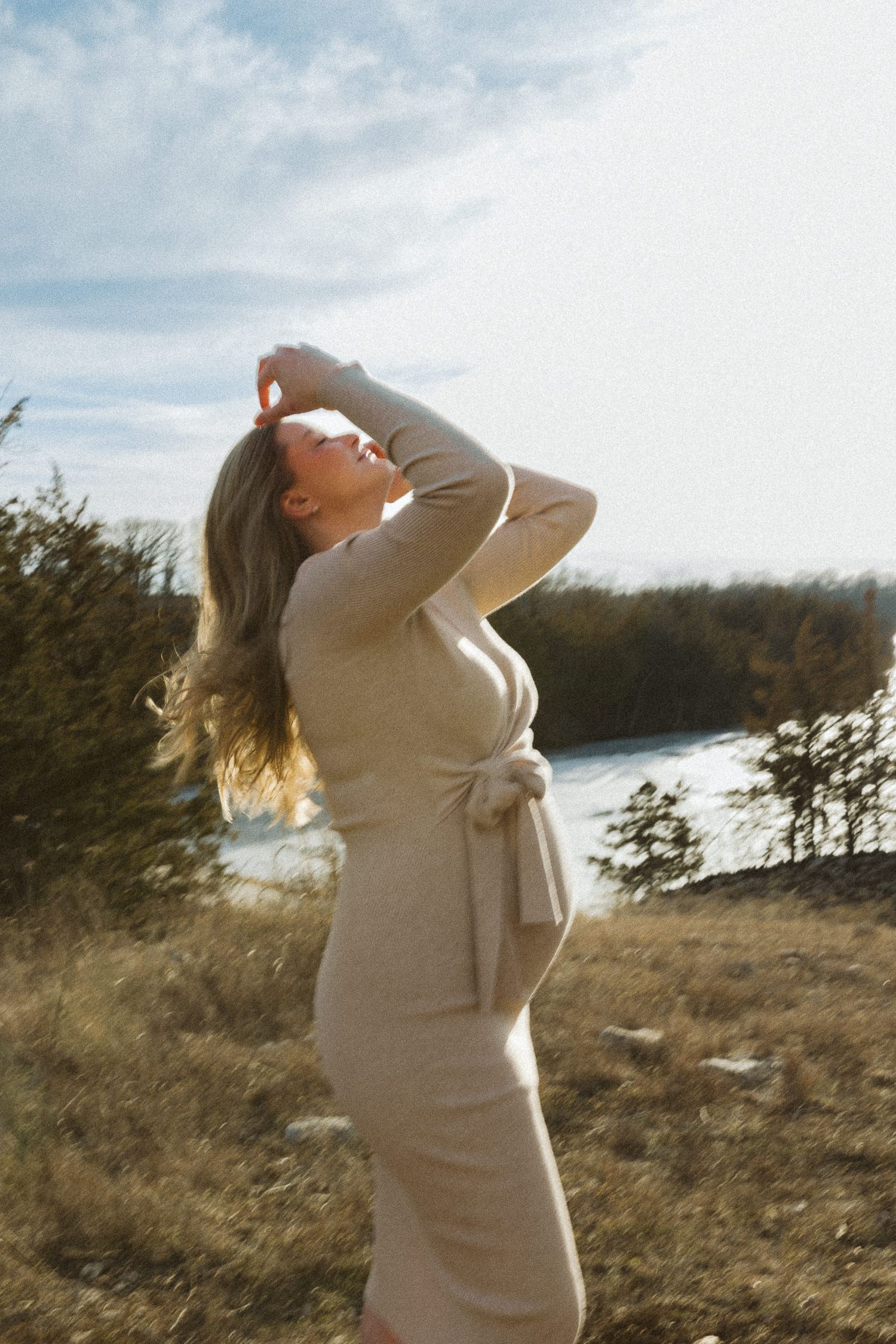 mom to be plays with her hair in the sunlight in front of pine tress with lake olathe behind her