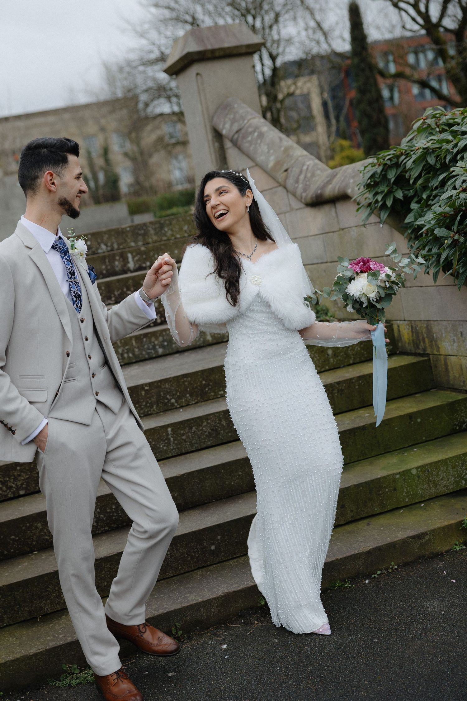 A bride in a white dress and groom in light gray suit dance playfully on stone steps during their wedding photoshoot.