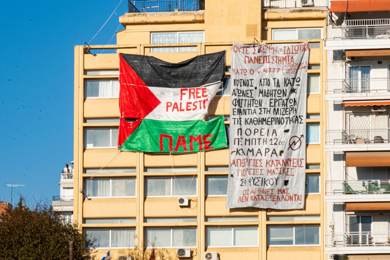 A Palestinian flag waves against a backdrop of apartment buildings with protest signs and banners visible.