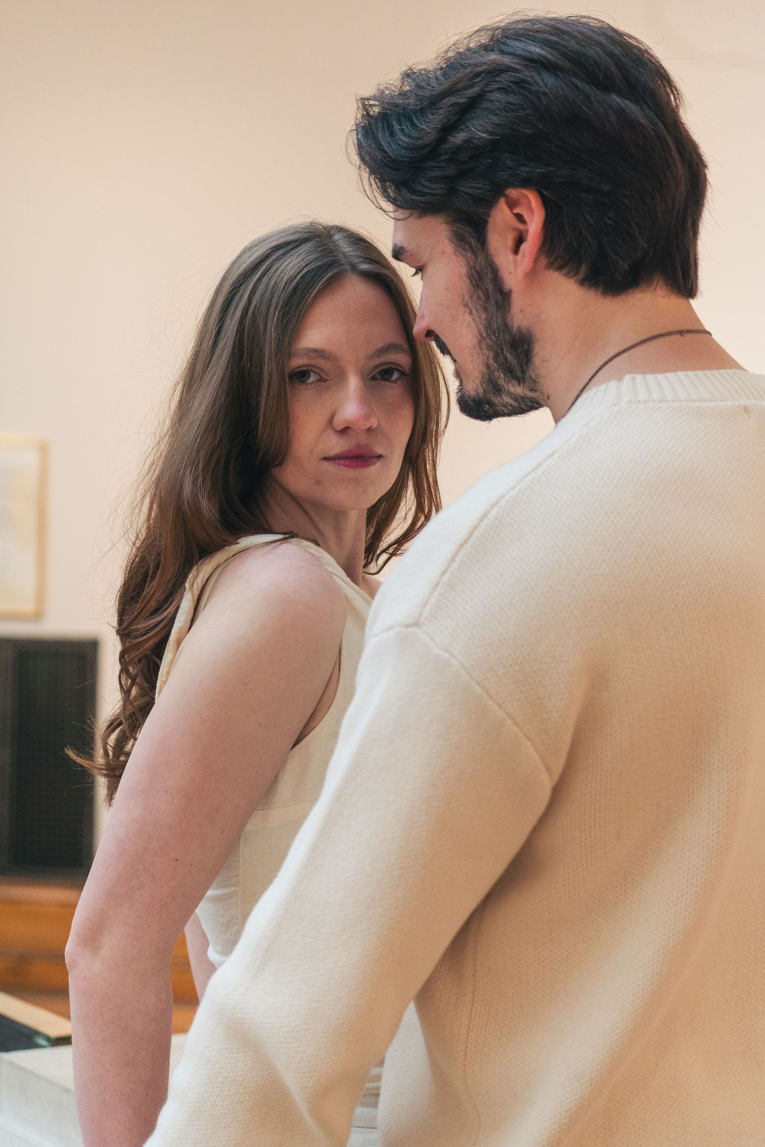 A romantic couple in cream outfits share an intimate moment, the woman gazing softly while the man leans close.