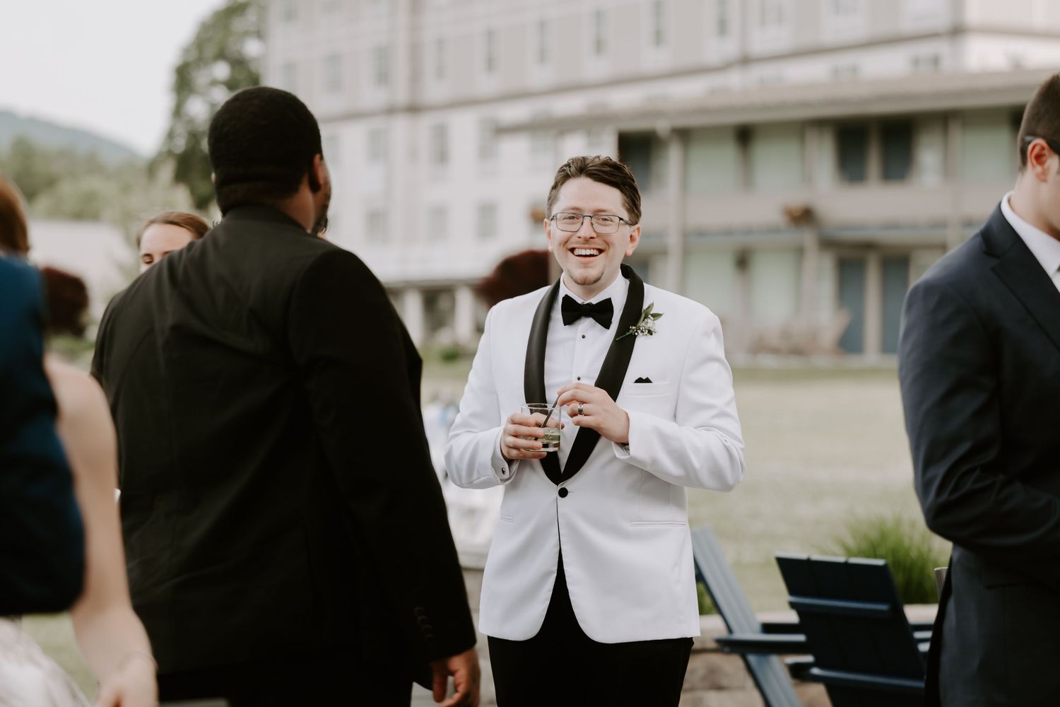 Smiling groom in white tuxedo jacket laughs with guests at outdoor wedding reception near historic building.