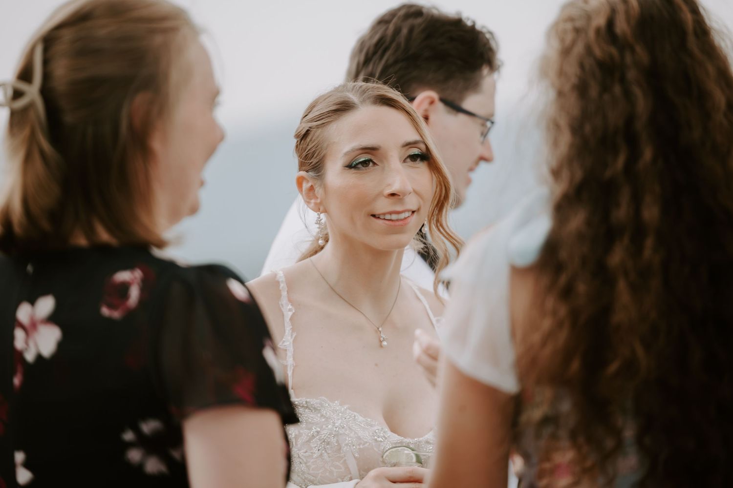 Bride in lace wedding gown smiles warmly while conversing with guests at outdoor wedding celebration.