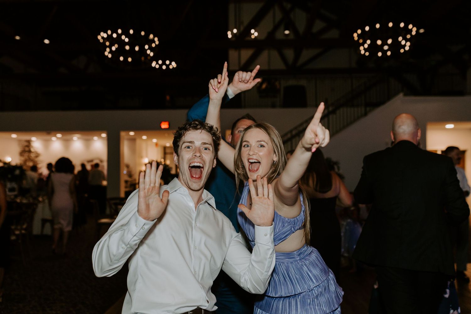 Two excited wedding guests celebrate on the dance floor with raised arms at lively wedding reception.