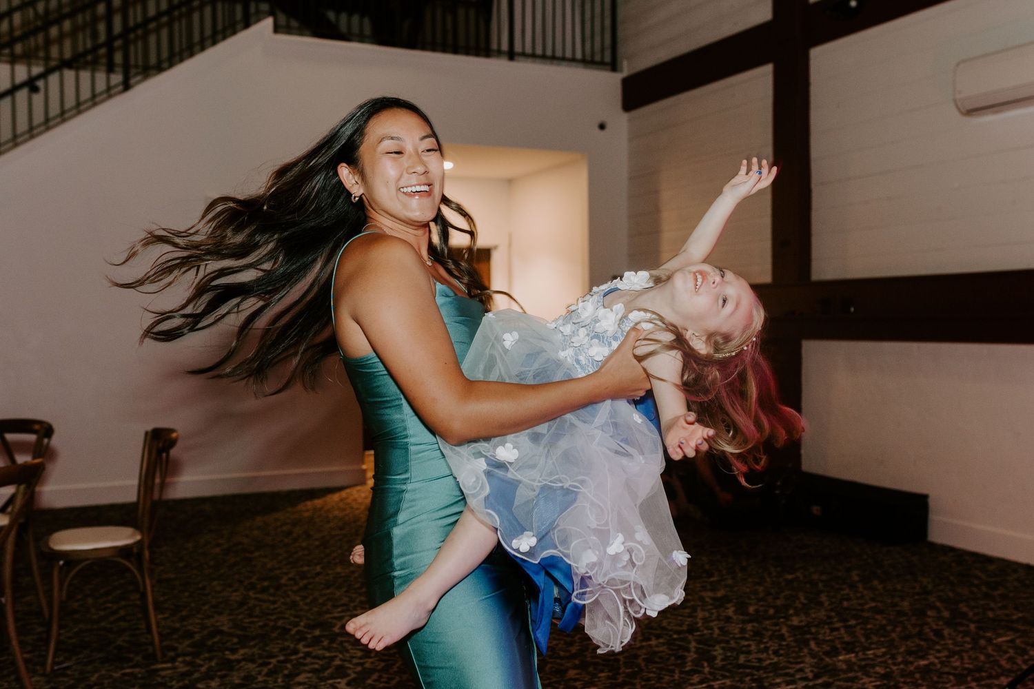 A woman in a teal dress joyfully lifts a young girl in a floral dress at a wedding reception.