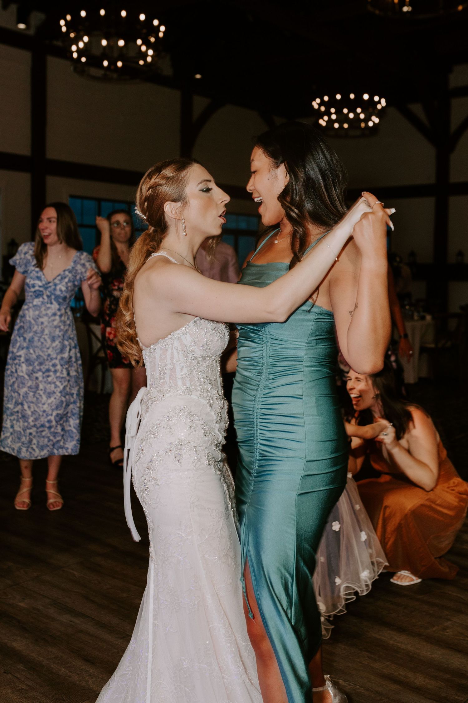 Bride in white gown and bridesmaid in teal satin dress dance together at wedding reception.