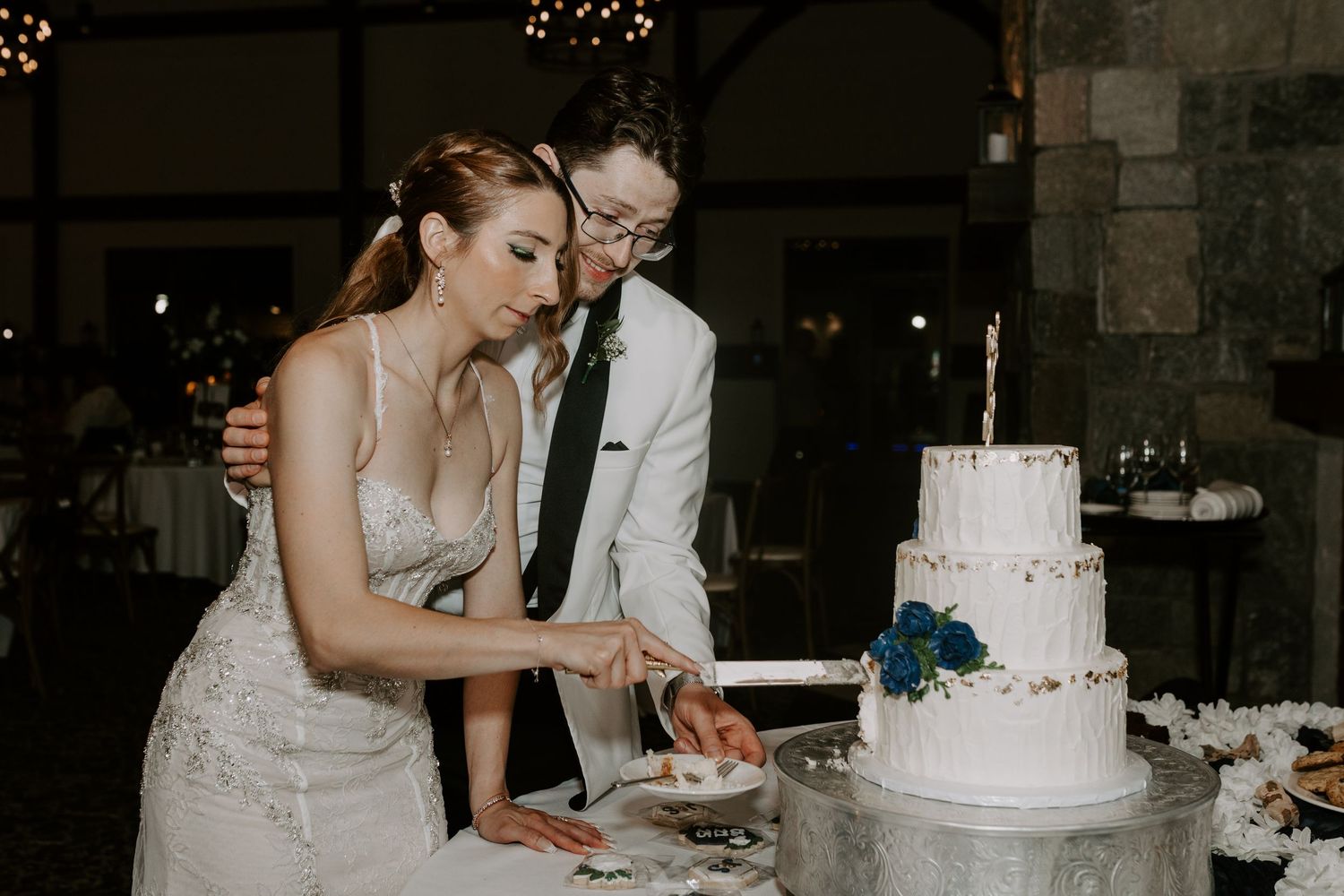 Bride and groom in white tuxedo cut their three-tier white wedding cake together.