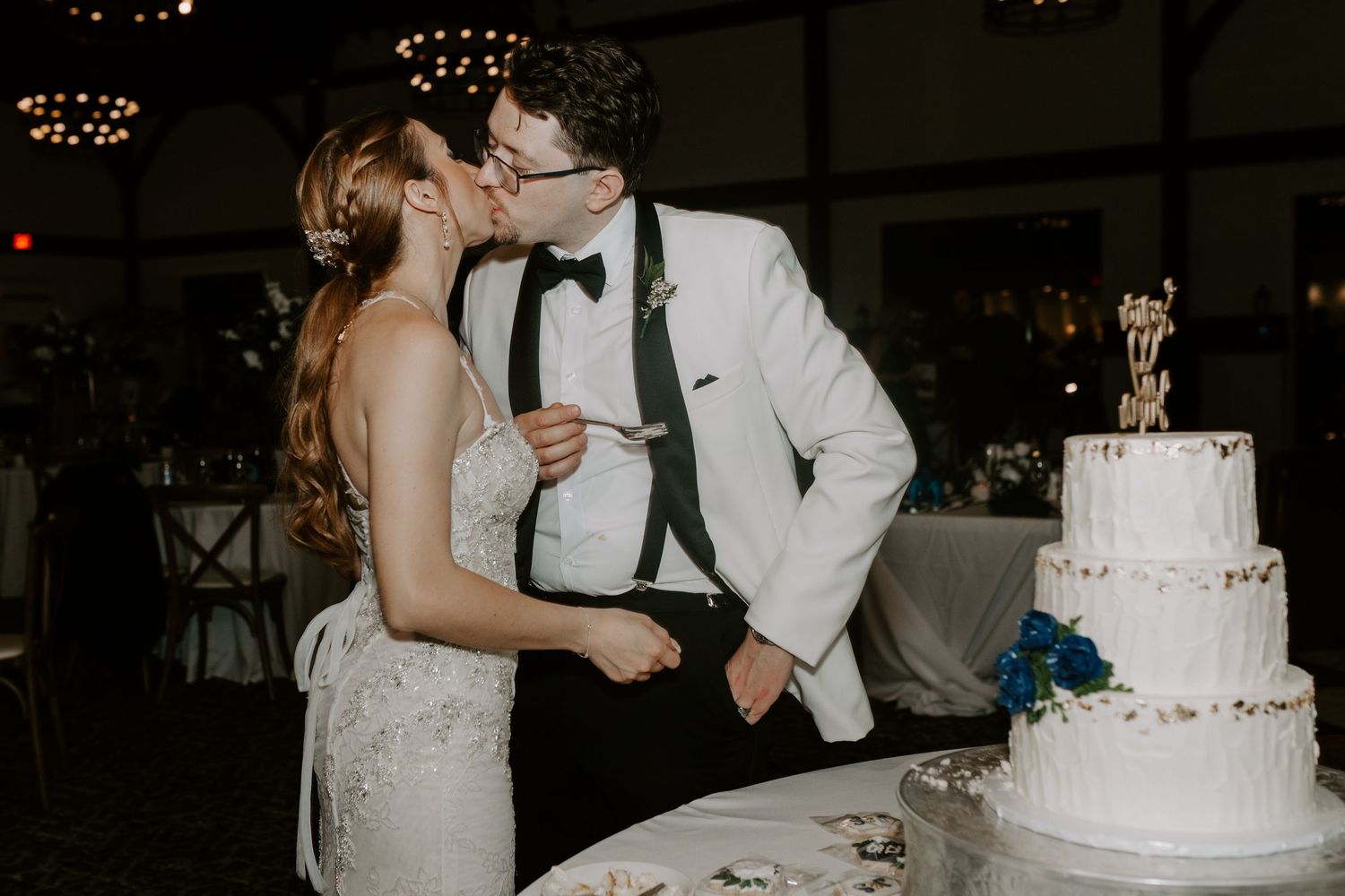 Bride and groom share a romantic kiss beside their decorated three-tier white wedding cake.