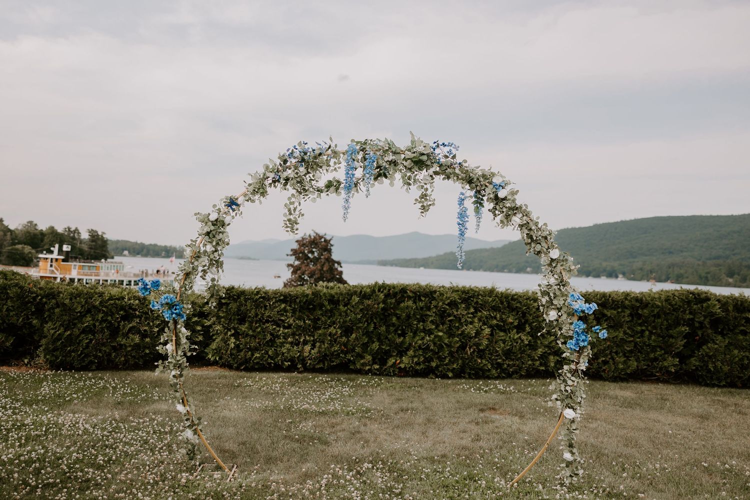 Circular floral wedding arch adorned with blue and white flowers overlooking a scenic lake and mountain view.