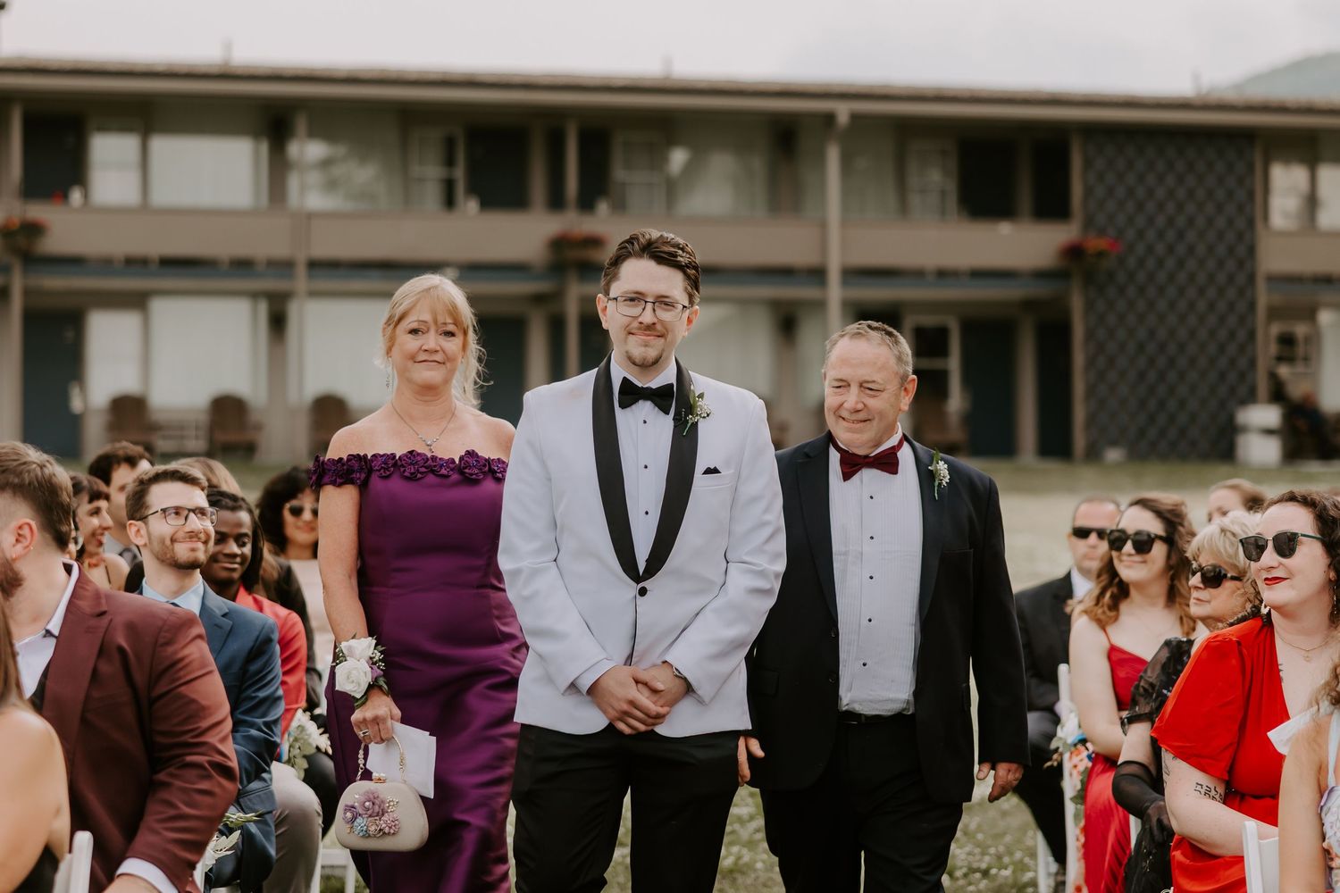 Groom in white tuxedo escorted down aisle by parents at outdoor wedding ceremony with seated guests.