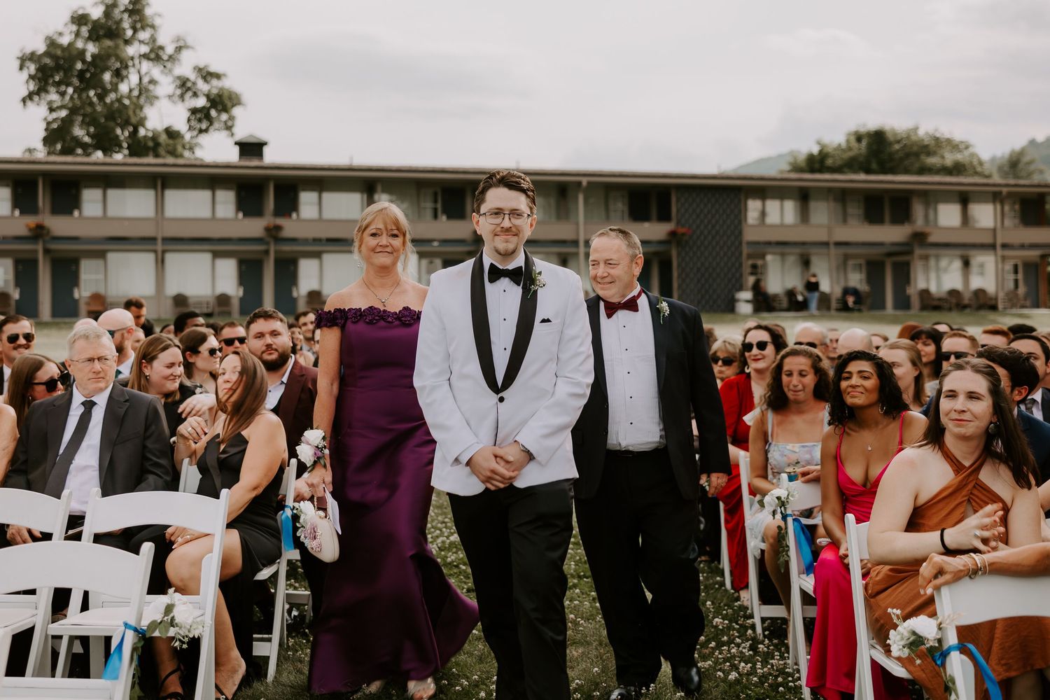 Groom in white tuxedo walks down aisle flanked by smiling parents at outdoor wedding ceremony.