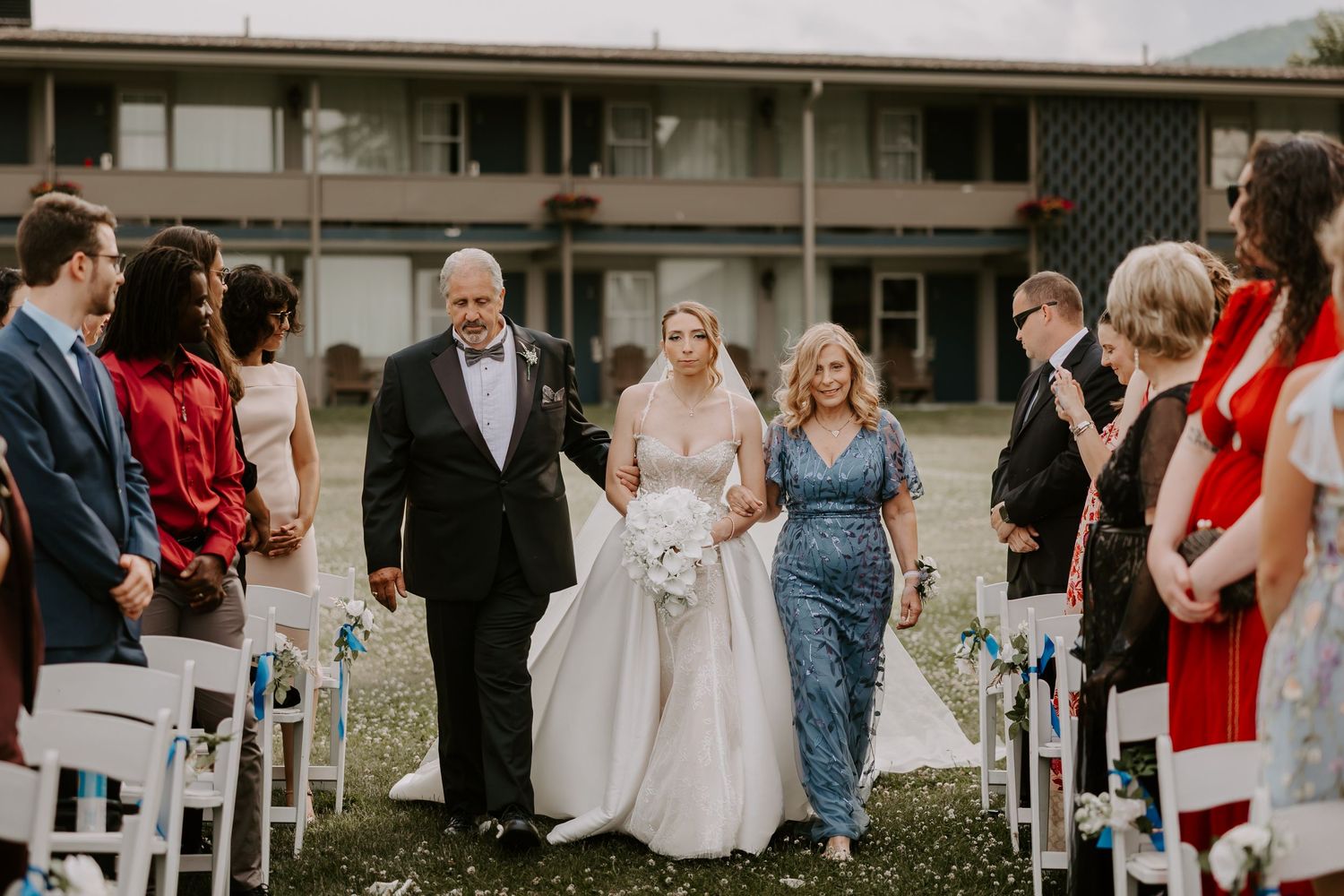 Bride in white gown walks down flower-lined aisle escorted by father and mother at outdoor ceremony.