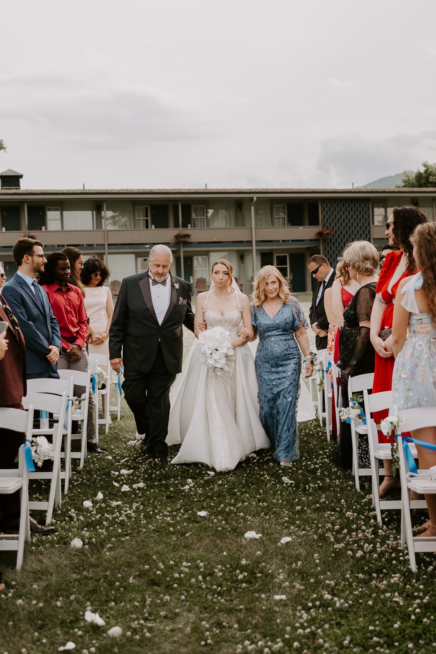 Bride in white gown and veil walks down aisle with parents at elegant outdoor wedding ceremony.