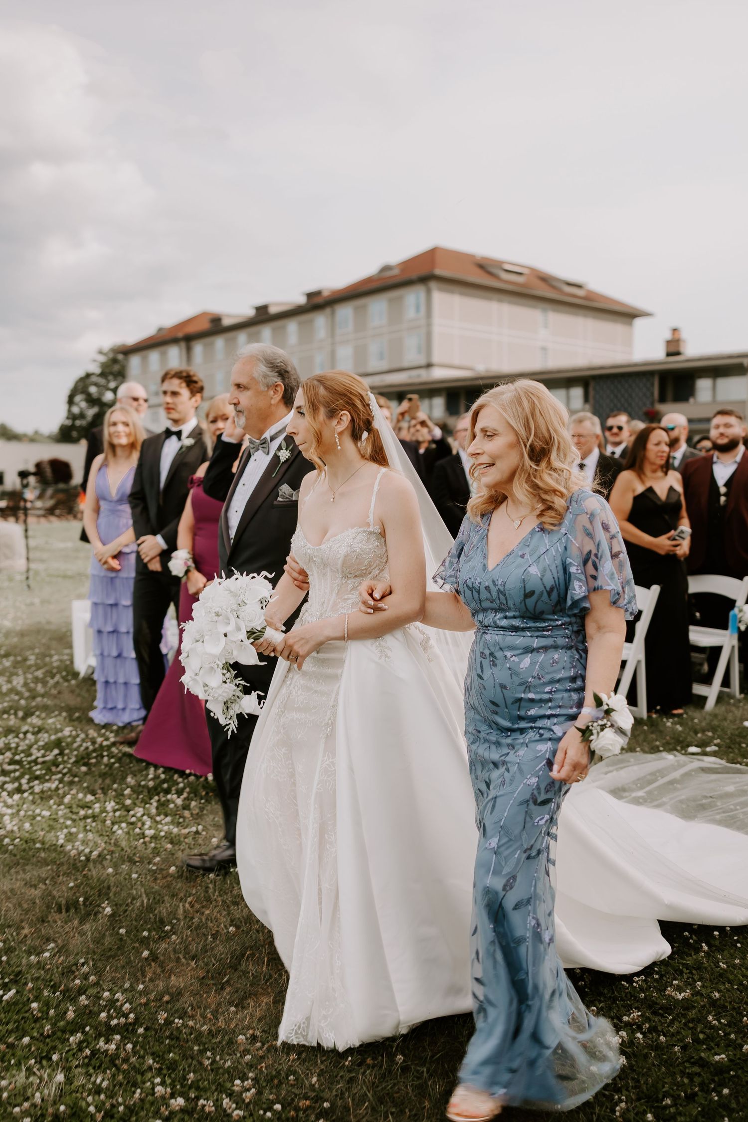 Bride in white gown walks down outdoor aisle with father in tuxedo and mother in blue floral dress.
