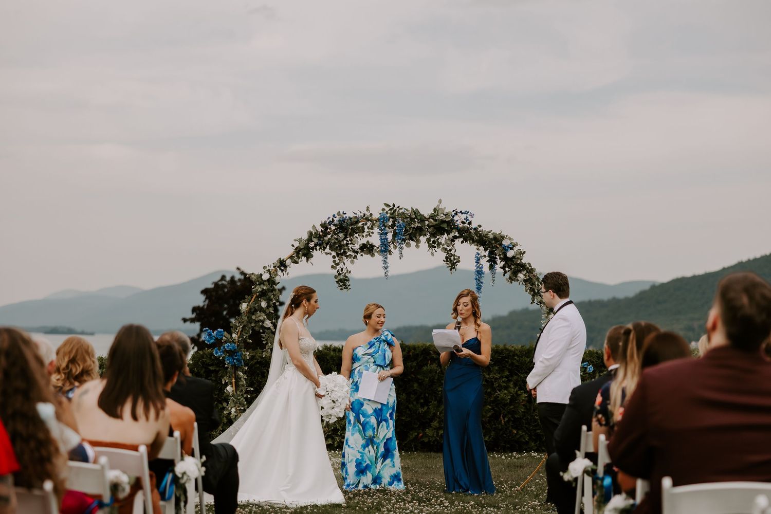 Couple exchanges vows under floral arch with mountain and lake views at outdoor wedding ceremony.