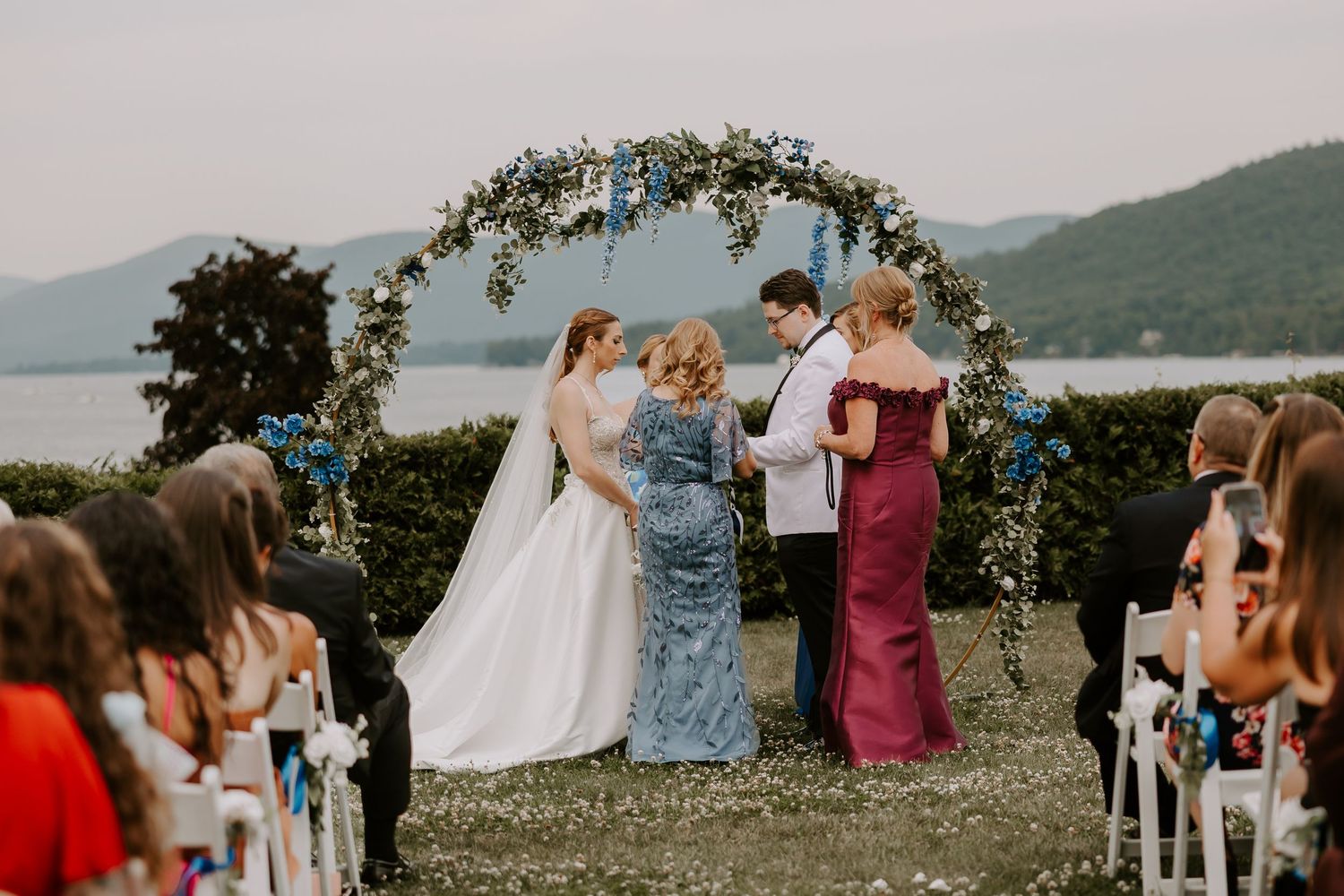 Wedding couple stands at floral circular arch with lake backdrop during outdoor ceremony with guests.