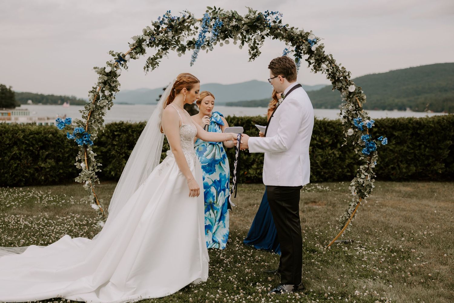 Bride and groom exchange vows under lush floral arch with mountain lake view at outdoor ceremony.