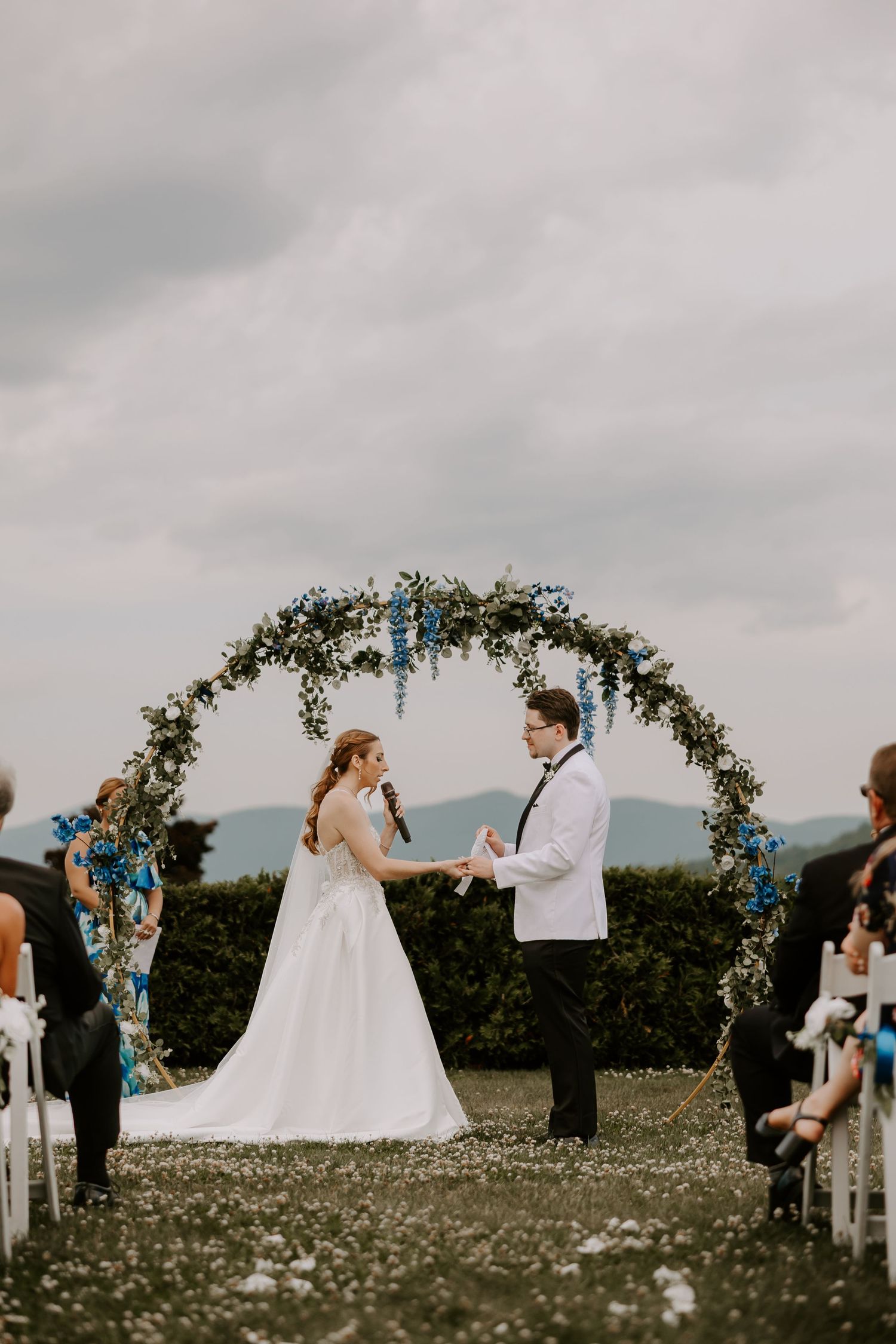 Bride and groom hold hands at outdoor wedding ceremony beneath circular floral arch with blue and white flowers.