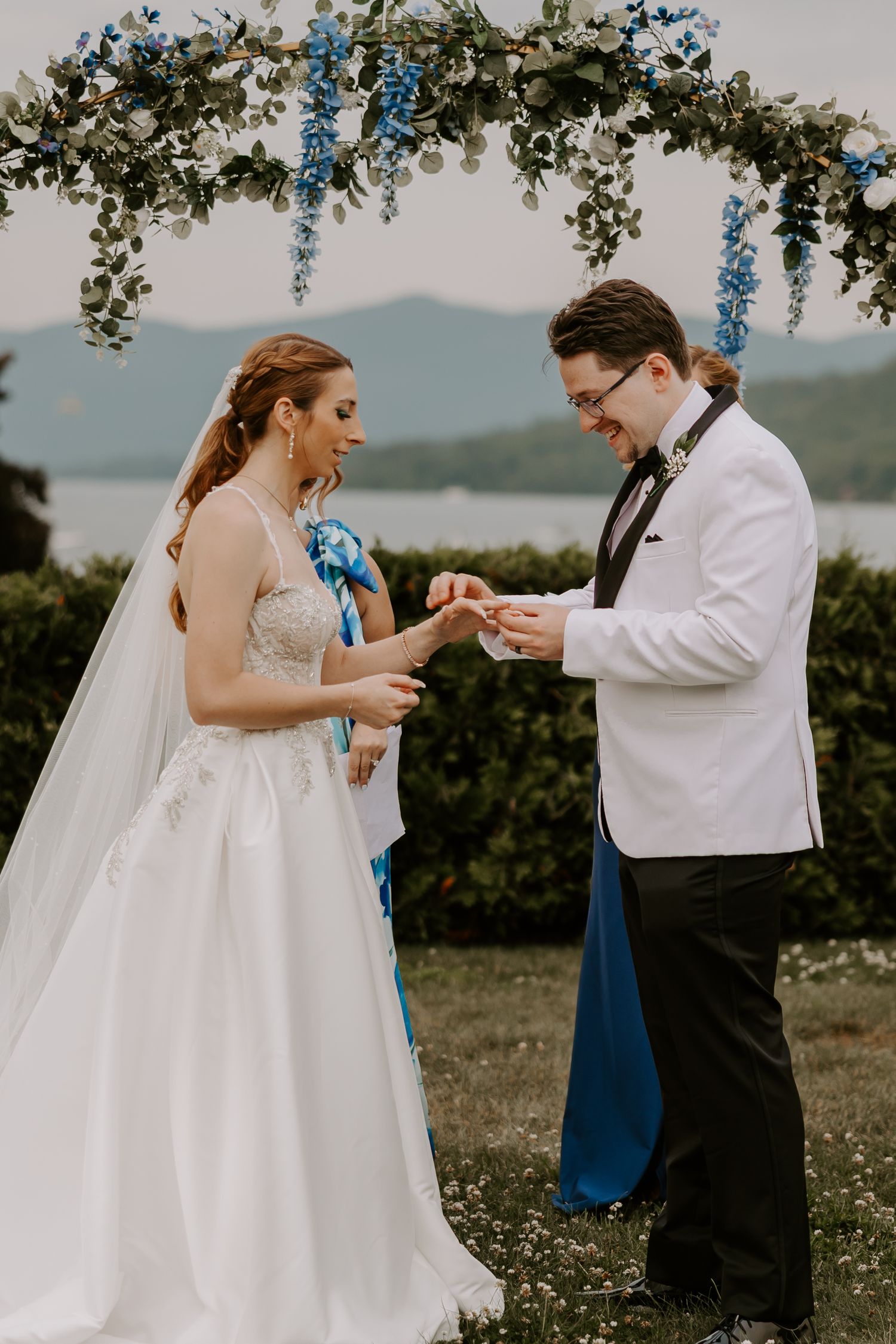 Groom places wedding ring on bride's finger under floral arch during outdoor ceremony with mountain backdrop.