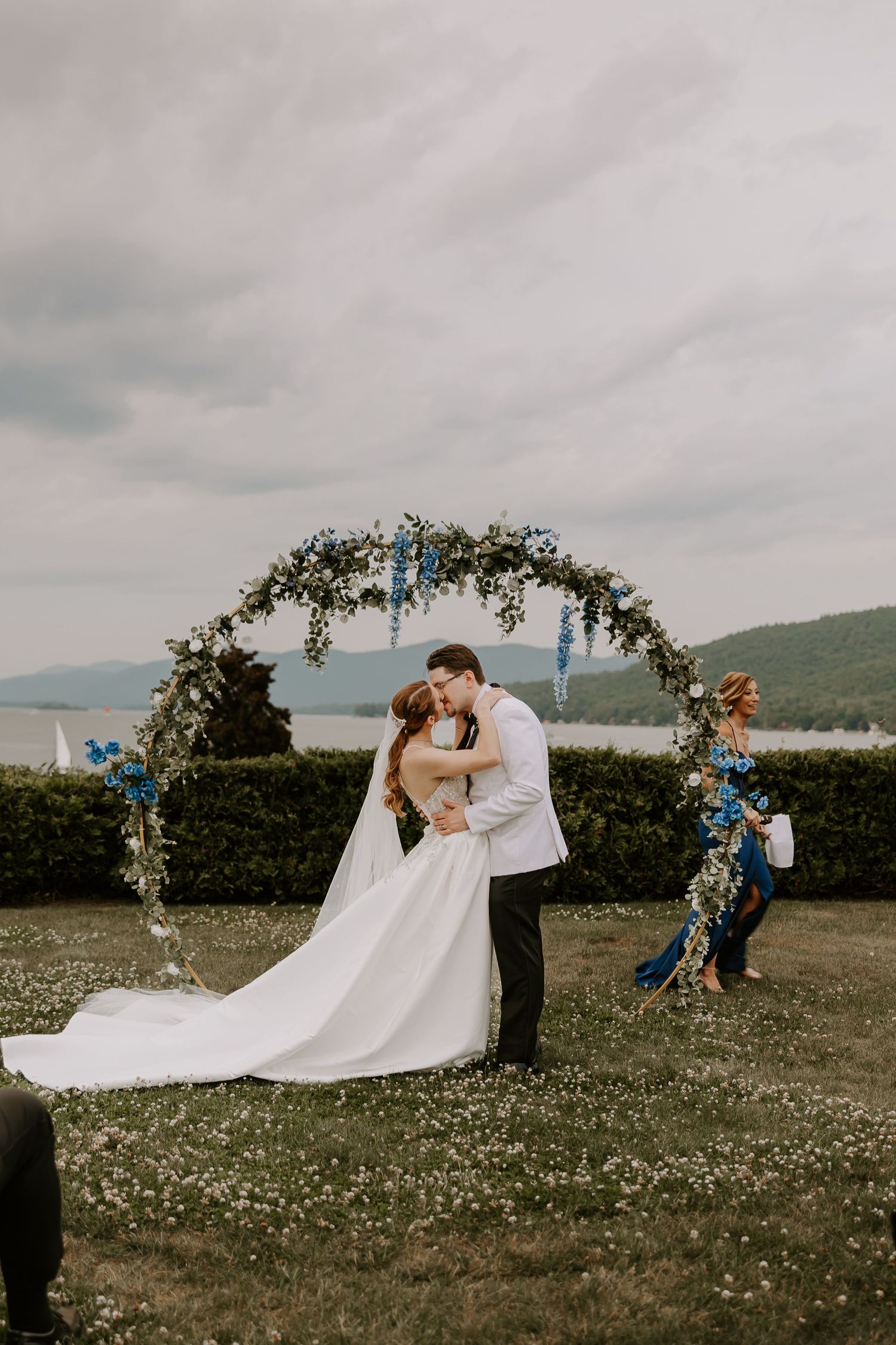 Newlyweds share first kiss embracing under circular floral arch at outdoor wedding with lake and mountain views.