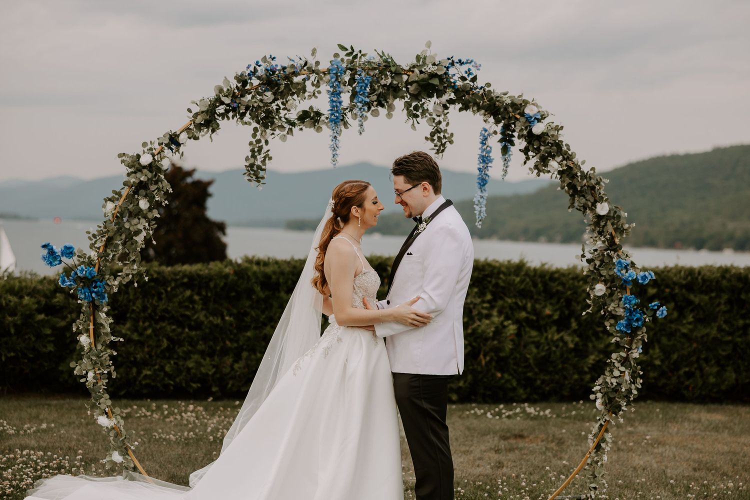 Bride and groom gaze at each other lovingly beneath circular greenery and blue floral arch at outdoor wedding.