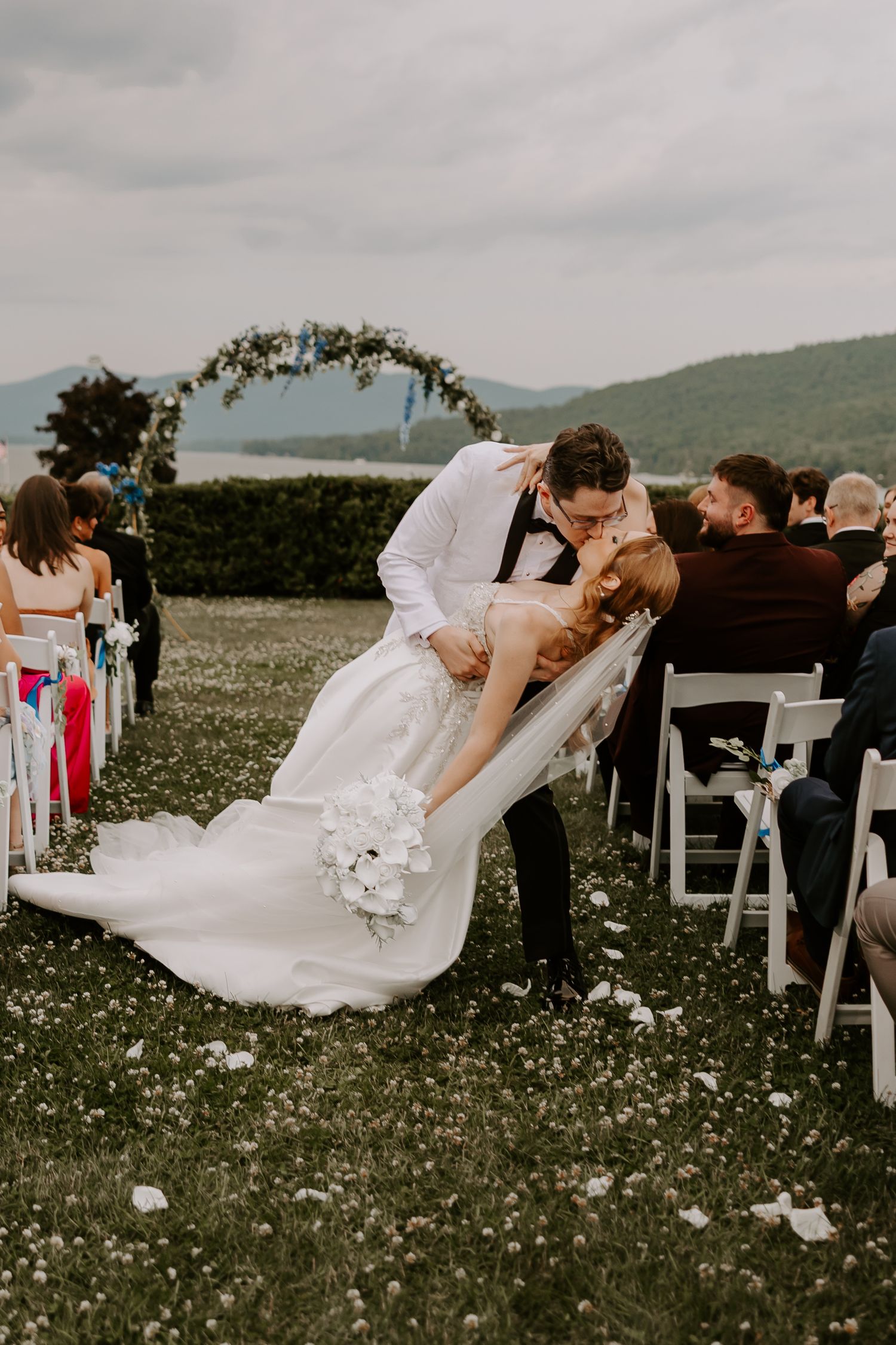 Groom dips bride for a romantic kiss in the aisle at outdoor wedding ceremony with floral arch backdrop.