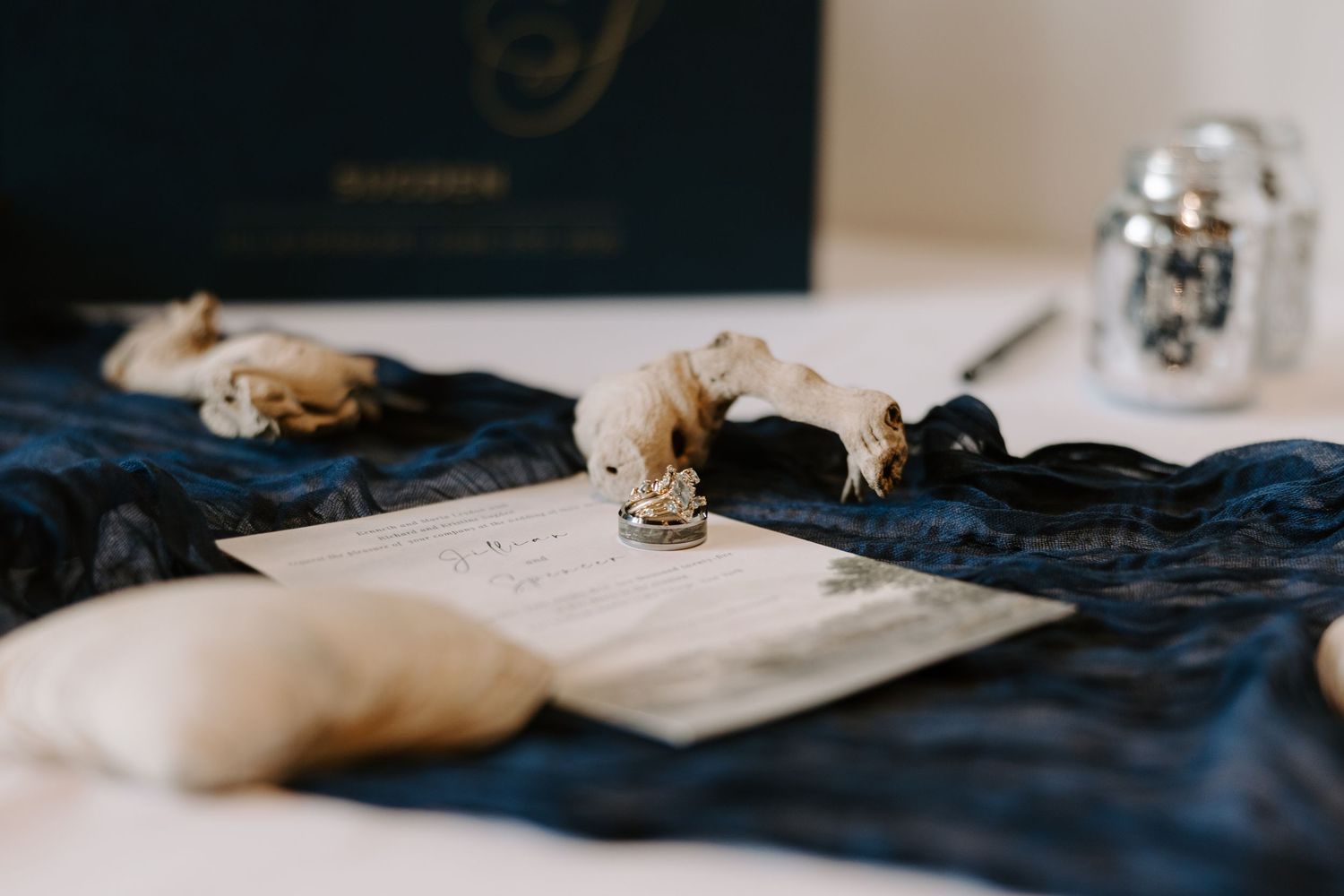 Wedding invitation suite with diamond ring displayed on navy blue silk fabric with decorative accents.