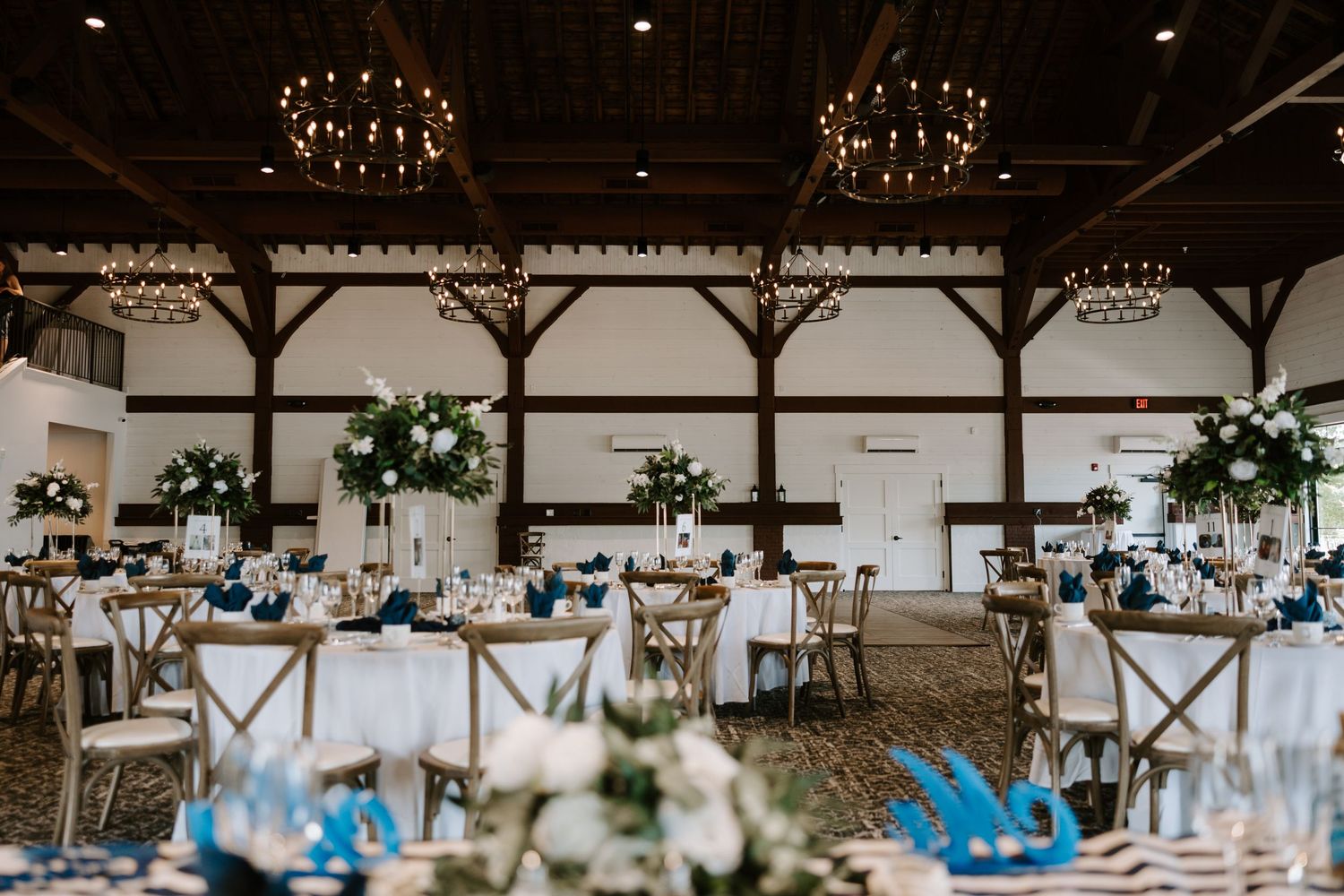 Elegant barn wedding reception hall with white floral centerpieces, blue accents, and chandelier lighting.