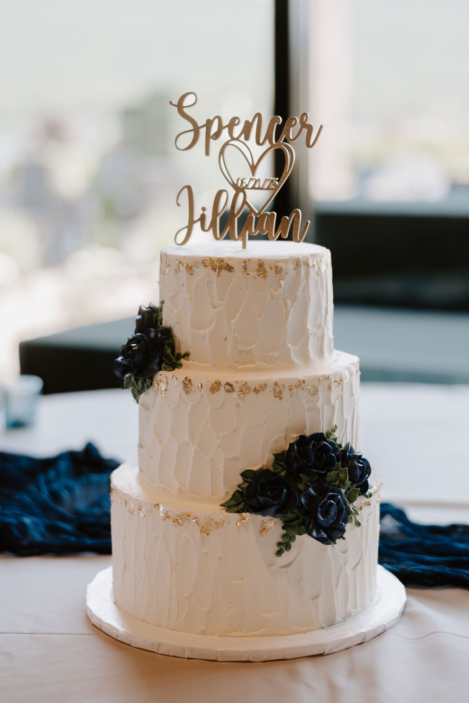 Three-tier white wedding cake with navy blue floral accents and custom Spencer and Jillian cake topper.