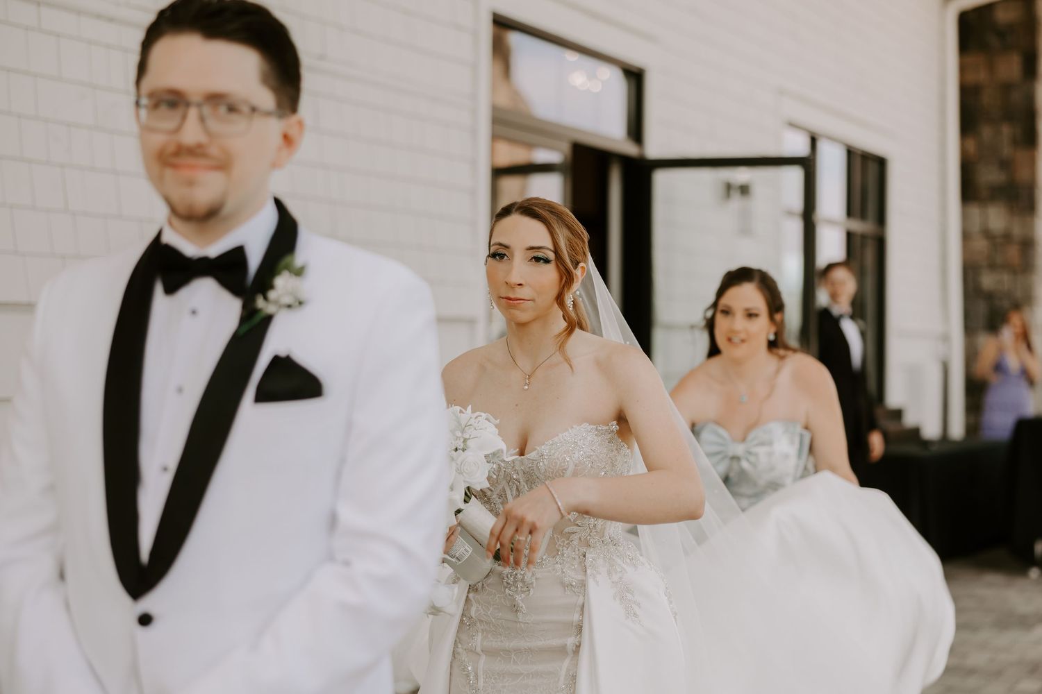 Bride in a strapless beaded gown approaches groom in white tuxedo for an outdoor first look moment.
