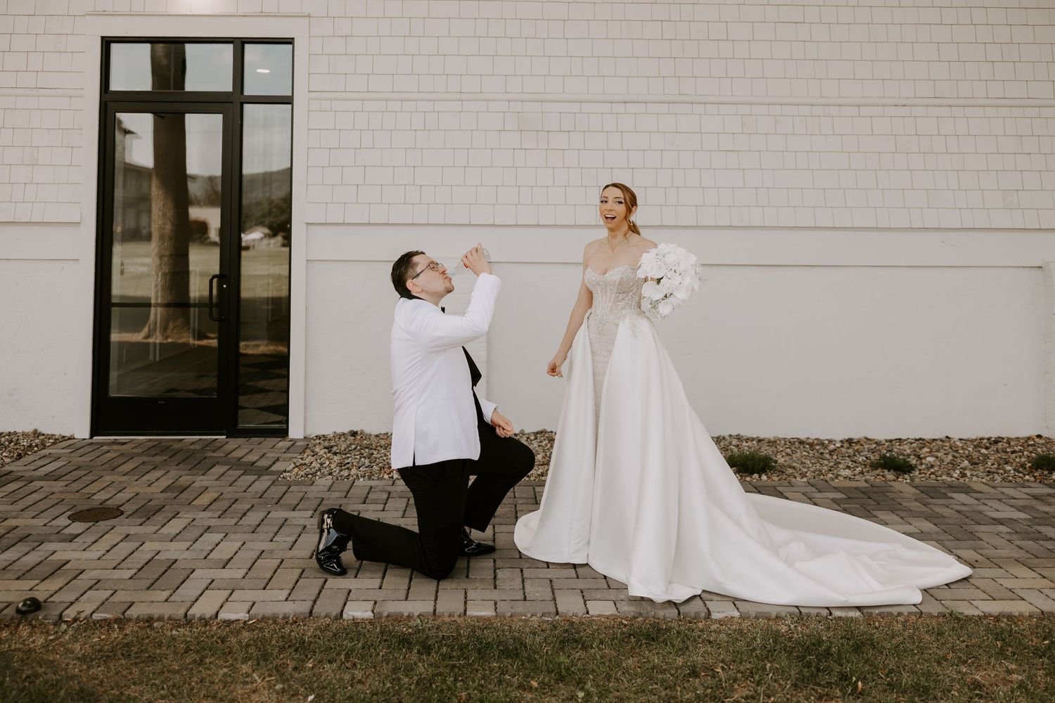 Groom dramatically kneels while bride laughs during a playful first look moment outside wedding venue.