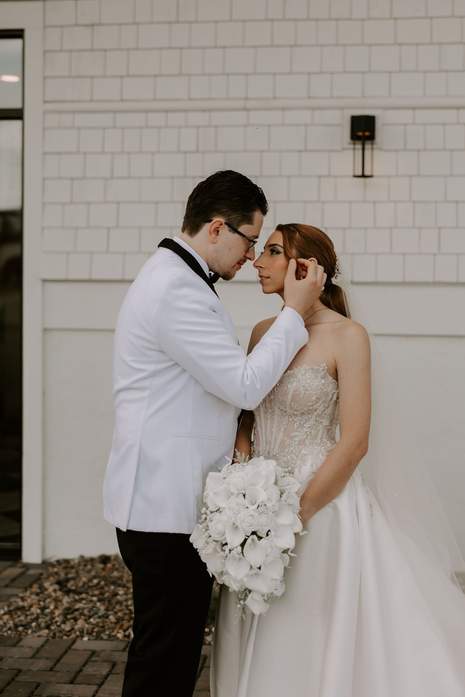 Bride and groom share an intimate forehead-to-forehead moment outside white shingle wedding venue wall.