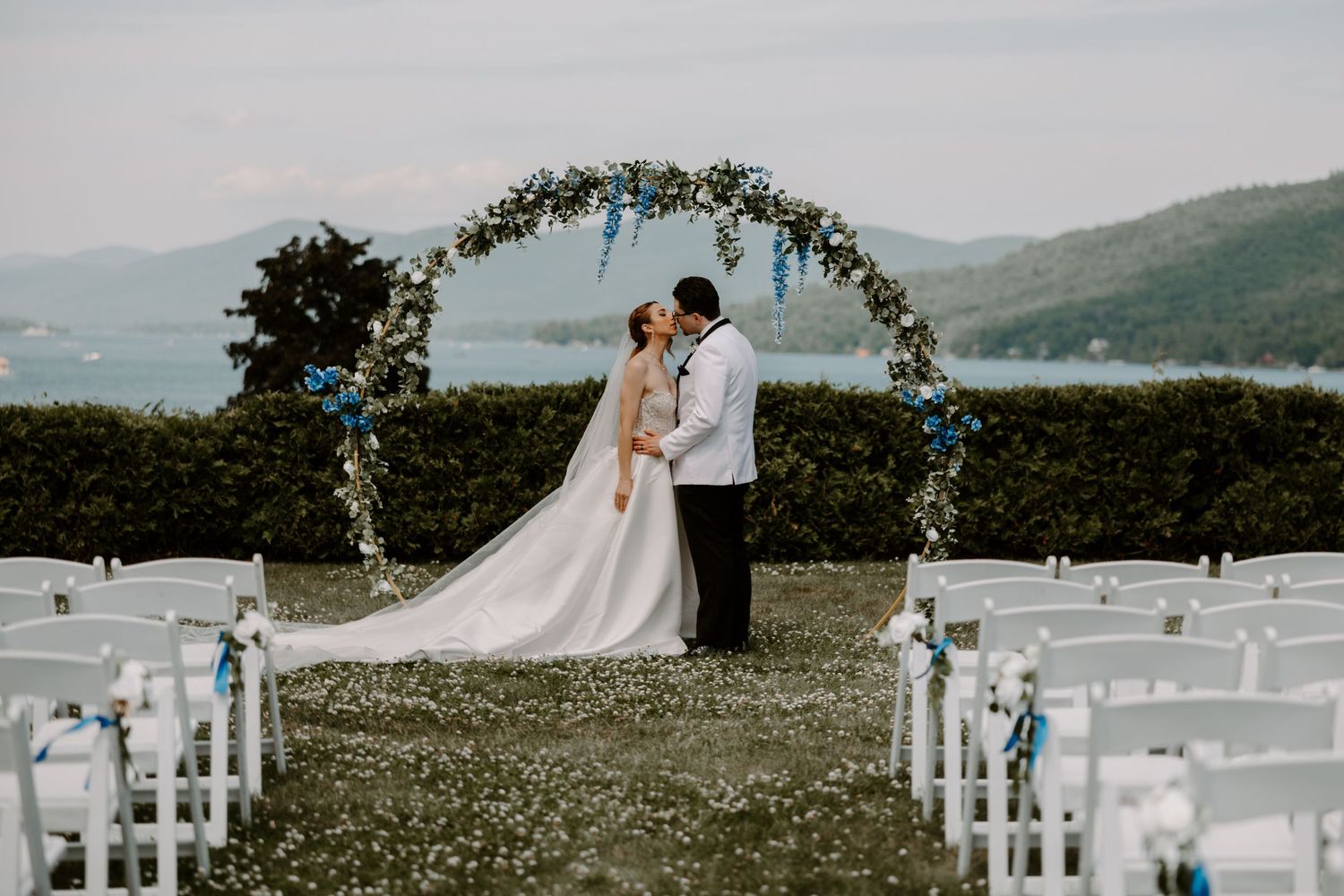 Bride and groom kissing under floral arch with blue flowers overlooking scenic lake view.