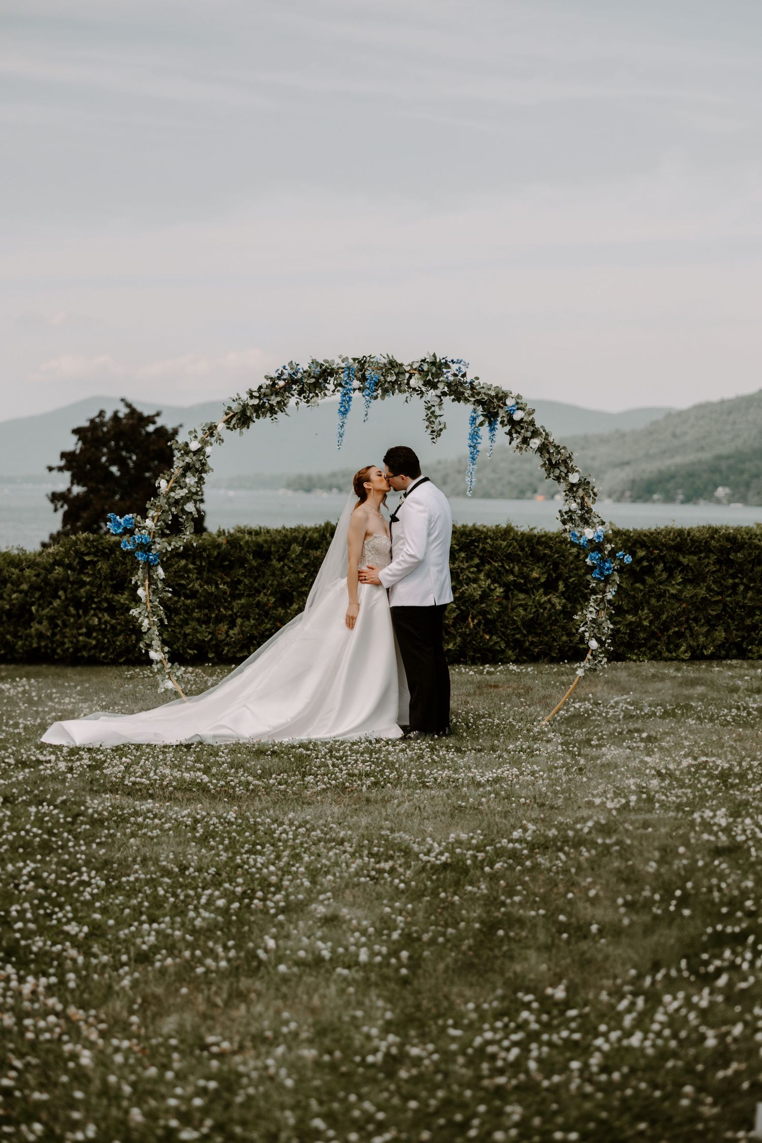 Couple kissing under circular floral arch with blue flowers in meadow with mountain backdrop.