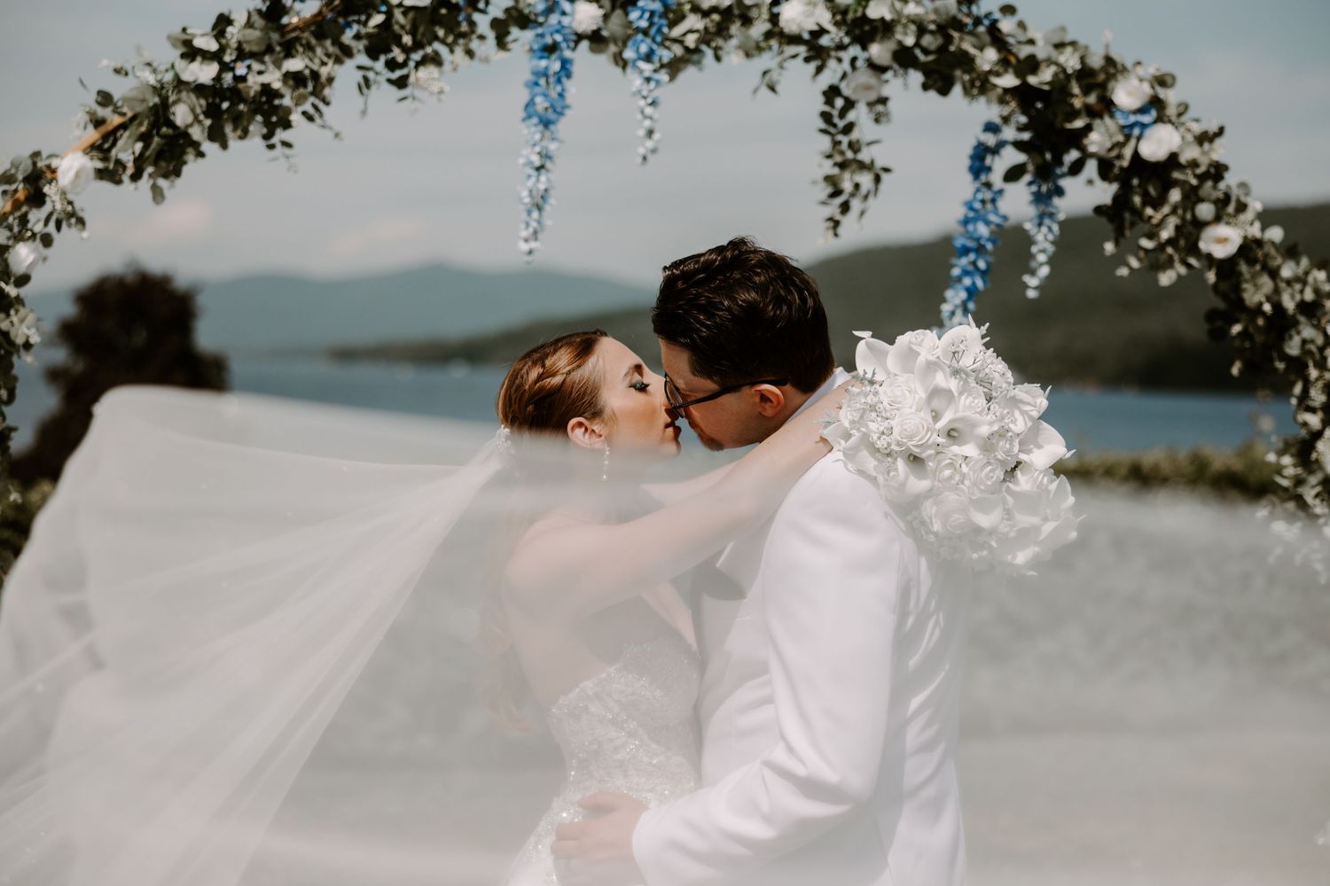 Bride and groom kissing under blue floral arch, veil flowing in wind over lake backdrop.