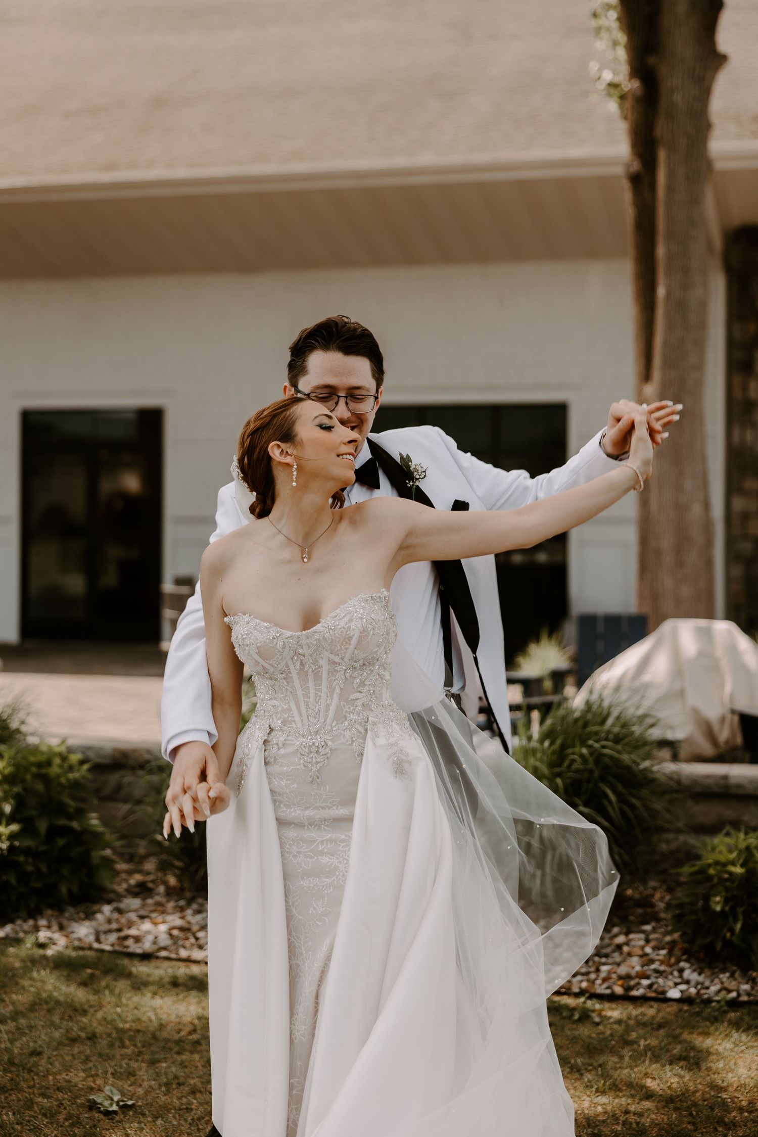 Groom in white jacket dancing with bride in lace wedding dress outdoors near building.