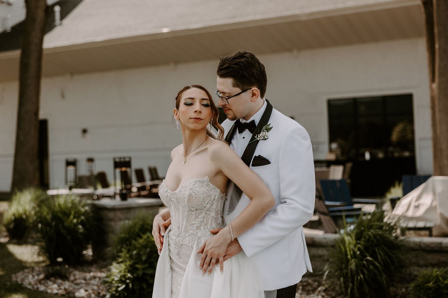 Bride in strapless lace gown embraced by groom in white tuxedo jacket in garden setting.