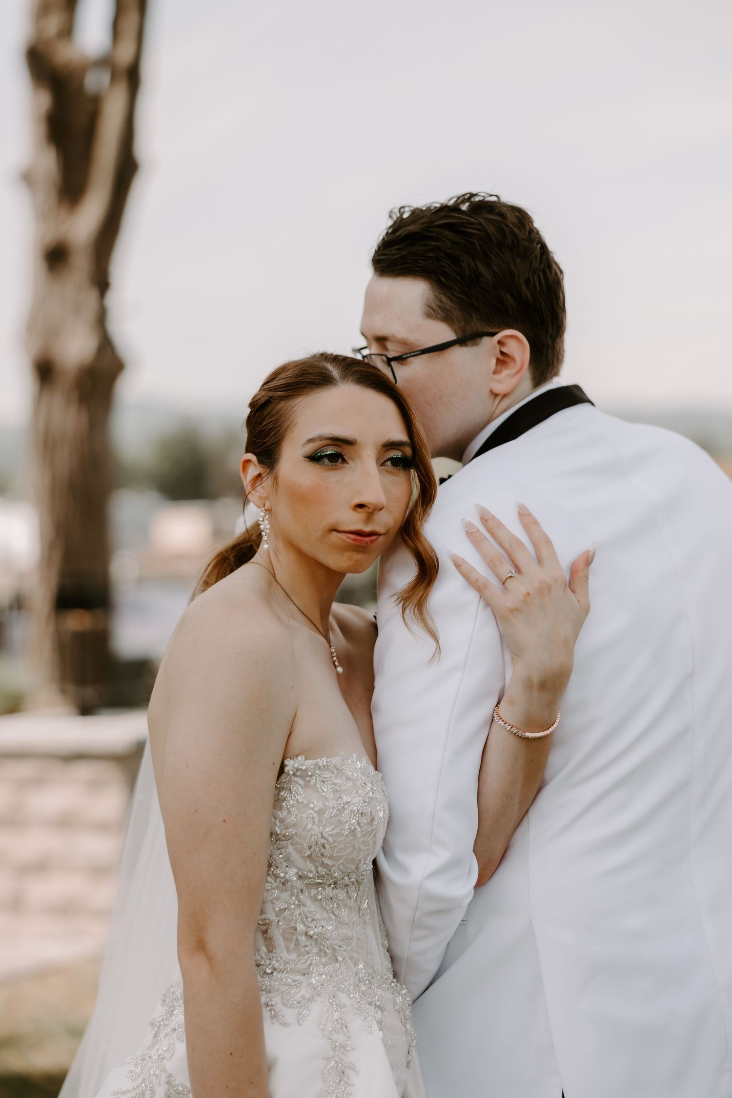 Groom in white tuxedo kissing bride on cheek while she gazes at camera outdoors.