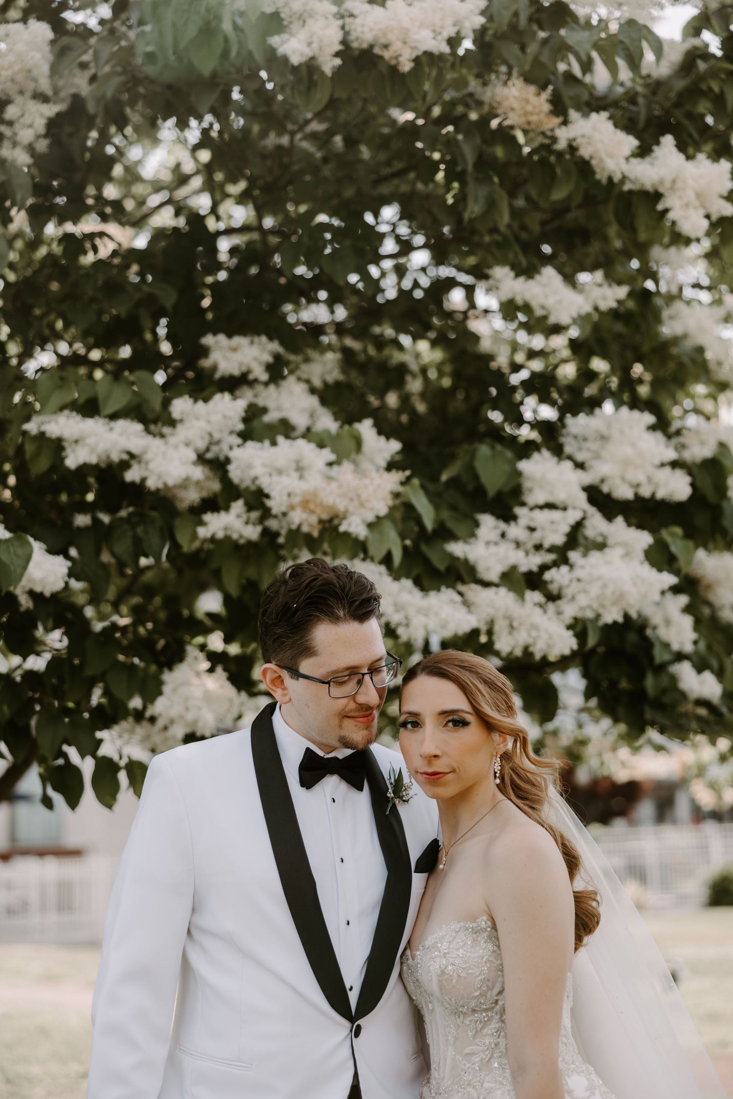 Bride and groom posing together under blooming white flowering tree in garden setting.