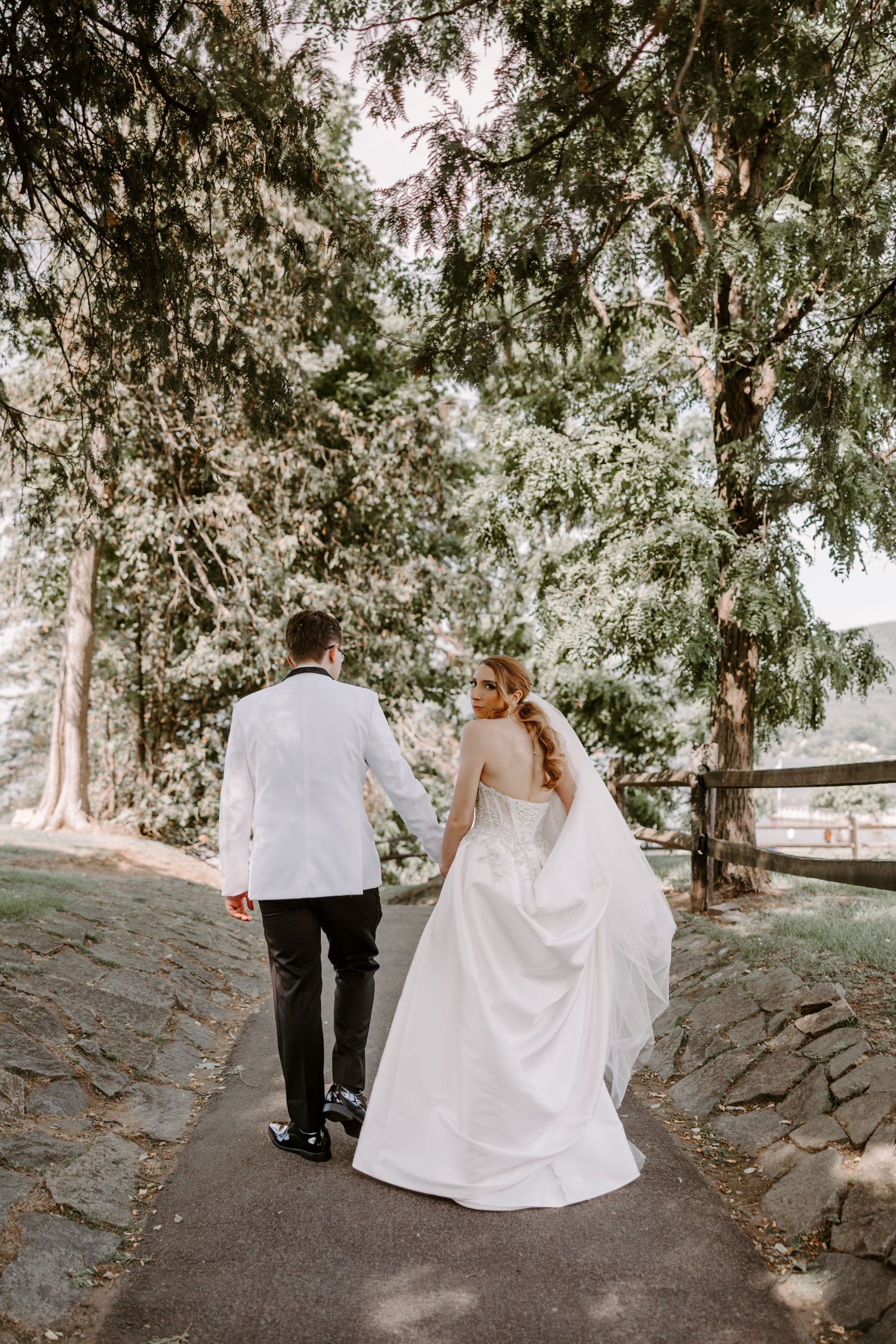 Bride and groom walking hand in hand along tree-lined path, viewed from behind.