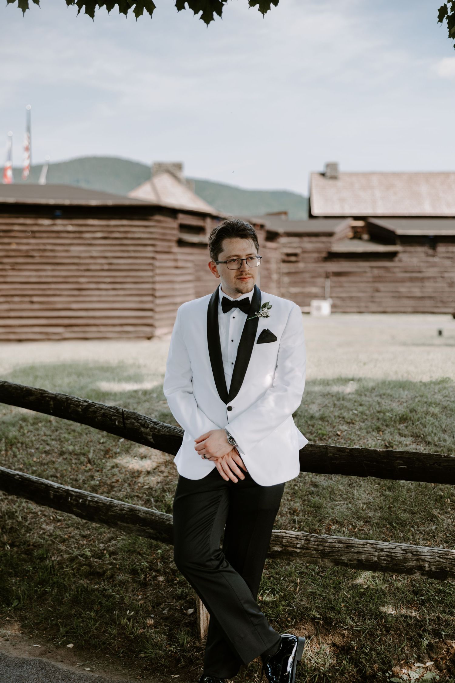 Groom in white tuxedo jacket and black bow tie leaning against rustic fence with mountain backdrop.