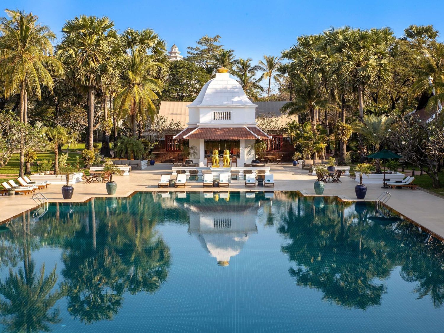 Luxurious resort swimming pool surrounded by palm trees and lounge chairs reflects in calm blue water at sunset.
