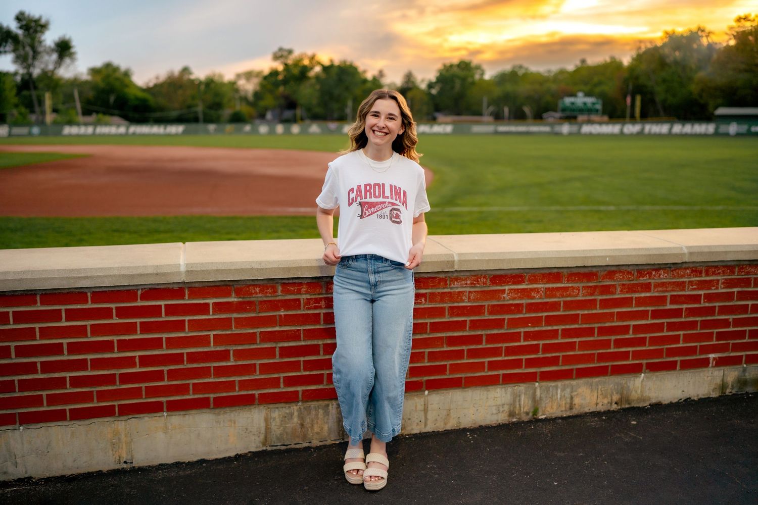 A person sits on a brick wall during sunset at an outdoor athletic field or stadium.