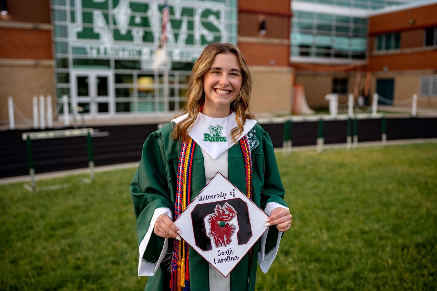 A graduate in green regalia holds up a decorated graduation cap outside a building with RAMS signage on display.